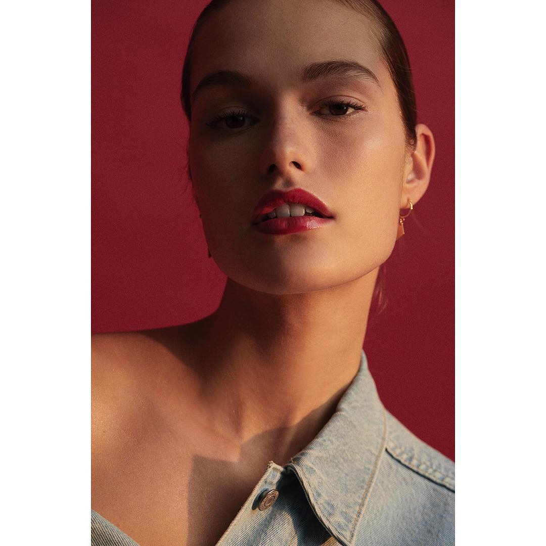 Close-up portrait of an adult female with red lipstick and a light denim jacket against a solid red background in a studio setting.