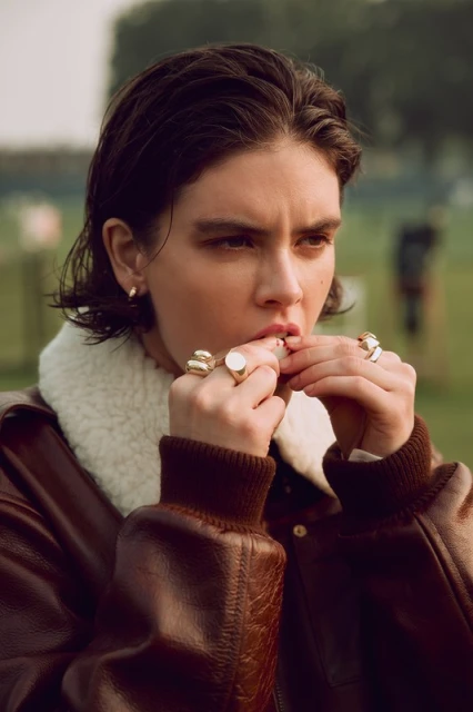 Portrait of a young adult with dark hair and wearing multiple rings, captured in an outdoor setting with soft, diffused light.