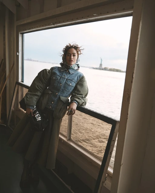 Woman in denim jacket and green skirt poses by a window with Statue of Liberty in the distance on a cloudy day.