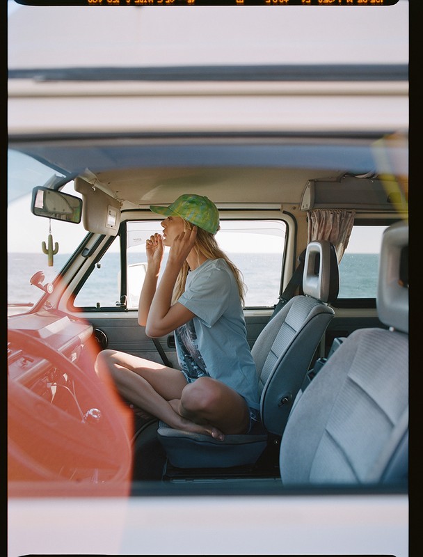 Woman in campervan wearing green cap, applying makeup near ocean, travel lifestyle, sunny day, light blue t-shirt, casual scene.