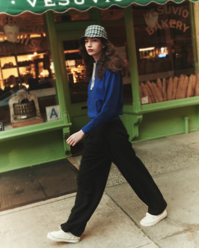 Young woman in blue hoodie walks past Vesuvio Bakery, wearing bucket hat, black trousers, and white sneakers on a city sidewalk.