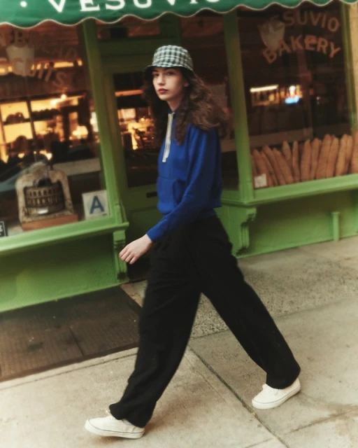 Young woman in blue hoodie walks past Vesuvio Bakery, wearing bucket hat, black trousers, and white sneakers on a city sidewalk.
