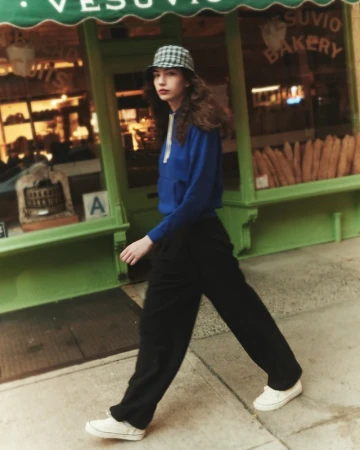 Young woman in blue hoodie walks past Vesuvio Bakery, wearing bucket hat, black trousers, and white sneakers on a city sidewalk.