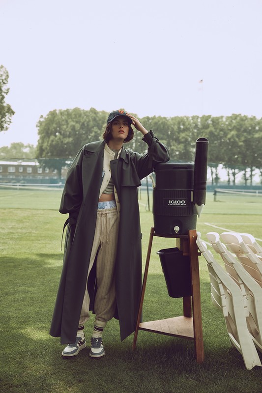 A fashionable adult woman stands next to an Igloo water cooler on a grassy lawn, wearing a trench coat and baseball cap in daylight.