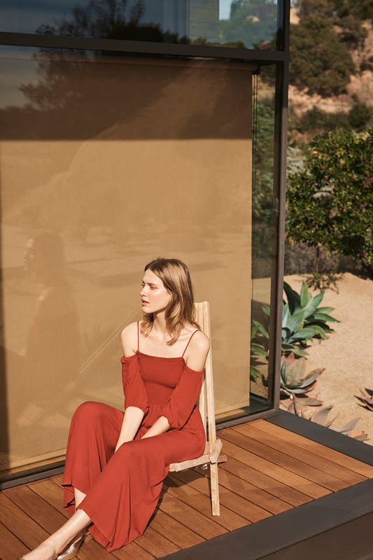 Adult woman in reddish-brown dress sitting on a wooden chair on a wood deck near a modern house with a neutral background.