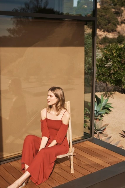 Adult woman in reddish-brown dress sitting on a wooden chair on a wood deck near a modern house with a neutral background.