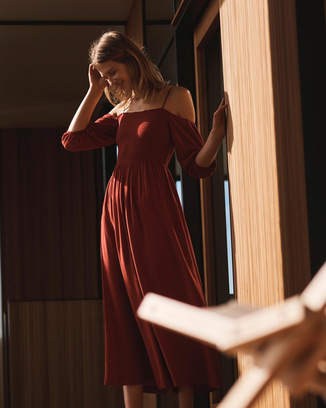 A stylish woman in a chic, rust-colored, off-the-shoulder dress, indoors with modern wood paneling, creating a warm and inviting fashion portrait.