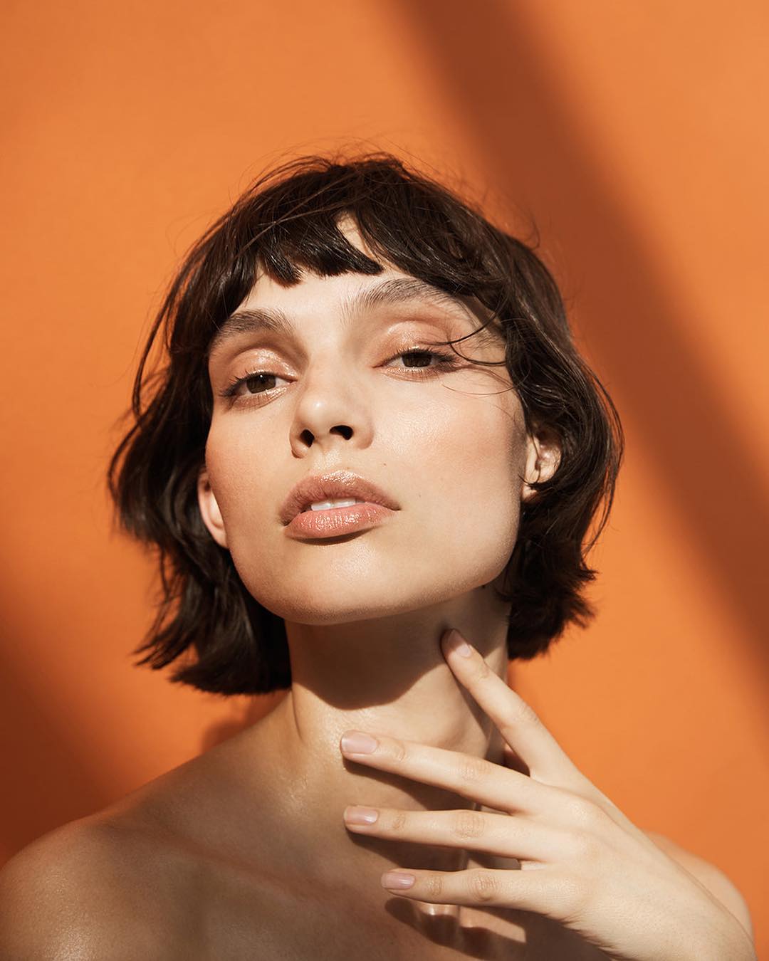 Close-up portrait of a woman with short dark hair and fair skin, hand on neck, against an orange backdrop with shadow pattern.