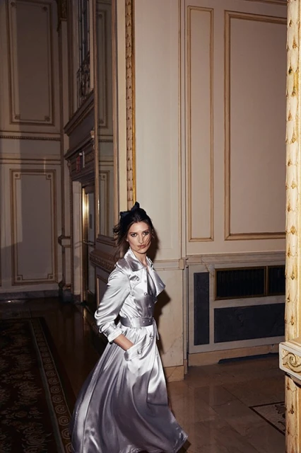A woman in a long satin coat, with a dramatic collar, poses in an ornately decorated hallway with high ceilings and gold trim.