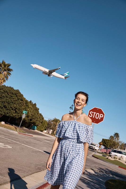 A woman in a blue and white dress stands on a street with an airplane in the background against a clear blue sky during the daytime.