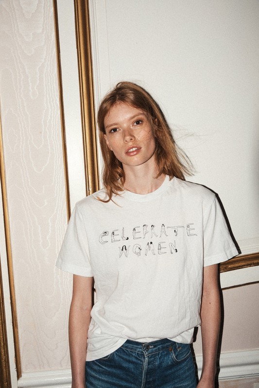 A redhead adult woman poses indoors wearing a white t-shirt with text and dark denim jeans looking at the camera in a studio setting.