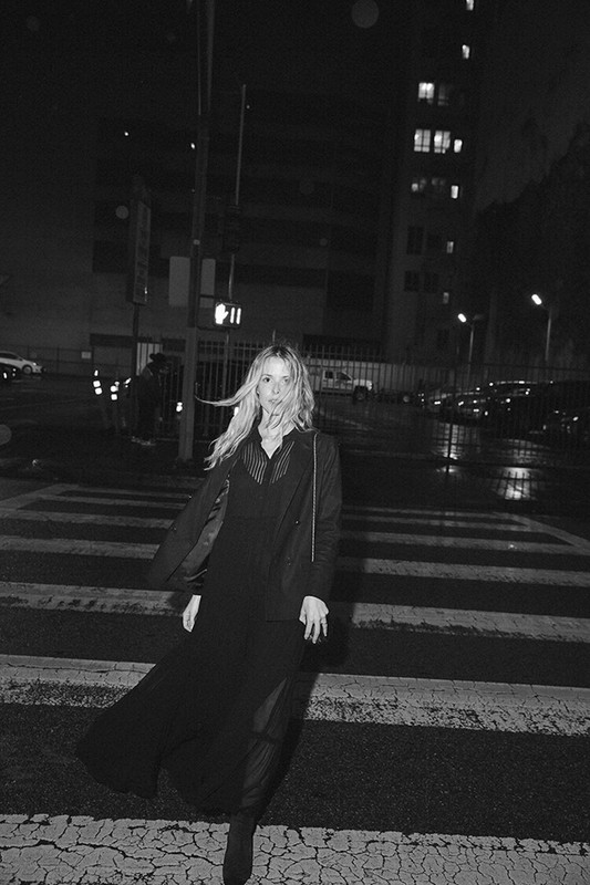 A woman with long blond hair poses on a crosswalk at night, wearing a long black dress and blazer, with city buildings in the background.