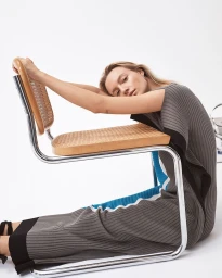 Woman in grey pleated dress resting arms on the wooden backrest of a tubular steel chair against a plain white background