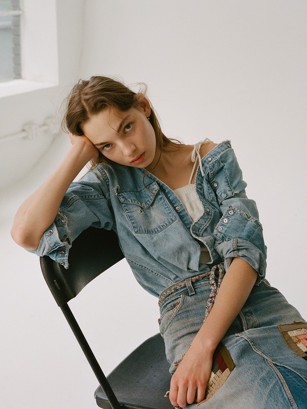 Young woman with long brown hair poses in a light blue denim jacket and jeans while seated on a black chair in a photo studio.