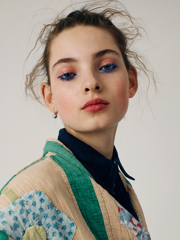 Portrait of a young woman with blue eyeliner and a patchwork jacket posing in front of a plain studio backdrop for a beauty or fashion shoot.