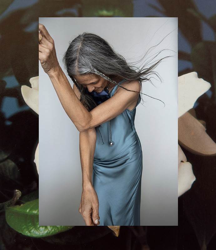 Elegant elderly woman with grey hair in a blue dress poses indoors with an out-of-focus floral background, creating a fine art fashion image.