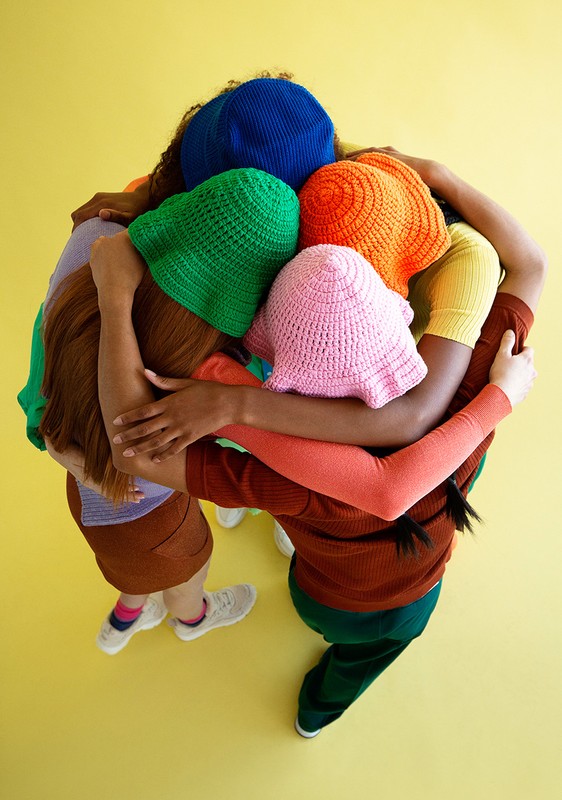 Group of young women wearing colorful knit hats huddle together on a yellow studio backdrop, viewed from an overhead angle, fashion, and unity.