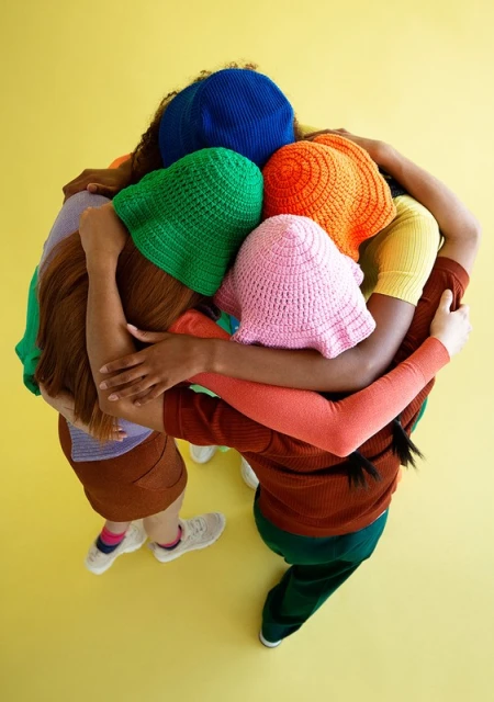 Group of young women wearing colorful knit hats huddle together on a yellow studio backdrop, viewed from an overhead angle, fashion, and unity.