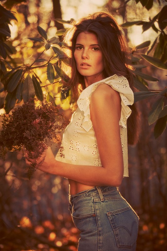 A young woman poses in a sun-drenched forest, holding foliage, wearing a white ruffled top and blue jeans in a fashion-style photograph.