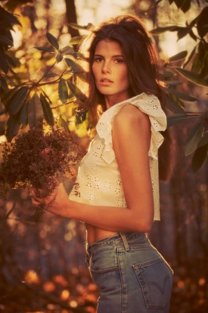 A young woman poses in a sun-drenched forest, holding foliage, wearing a white ruffled top and blue jeans in a fashion-style photograph.