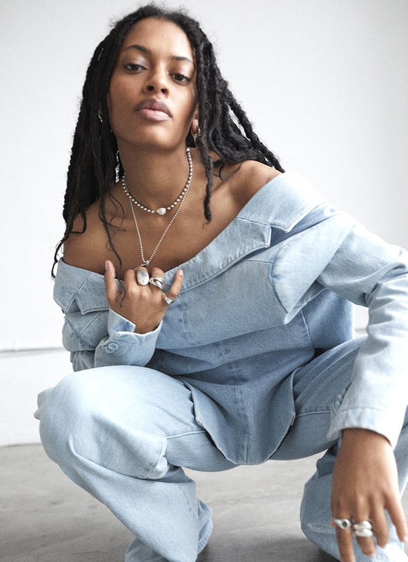 Young woman with dreadlocks poses in light denim off-shoulder top and pants, accessorized with jewelry against a neutral backdrop.