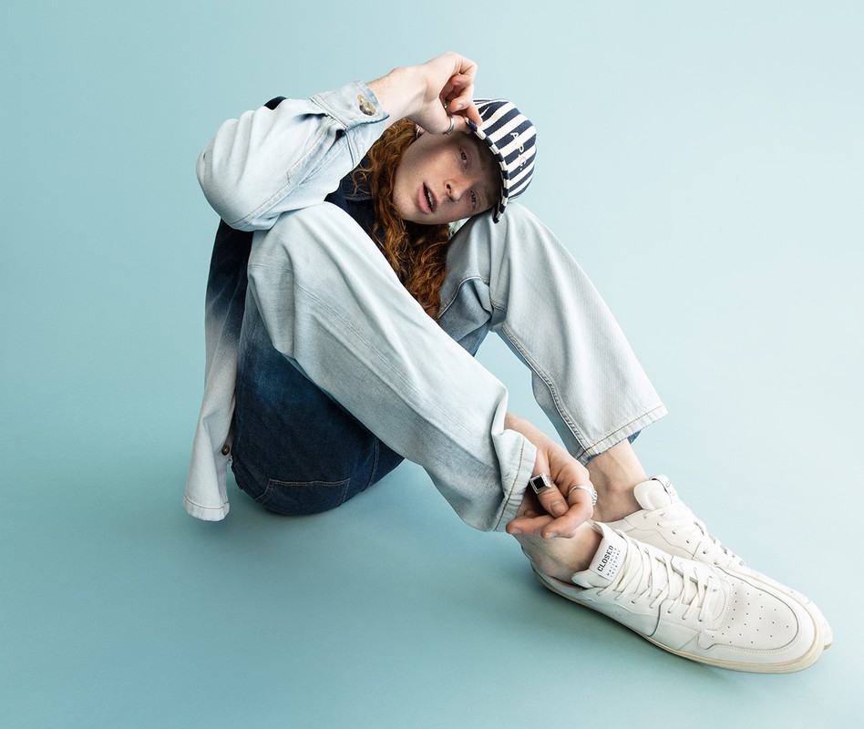 Young adult with red hair sits on a light blue background wearing a denim jacket, striped cap, jeans, and white sneakers.