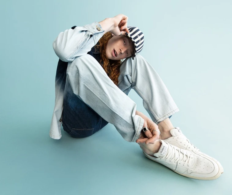 Young adult with red hair sits on a light blue background wearing a denim jacket, striped cap, jeans, and white sneakers.