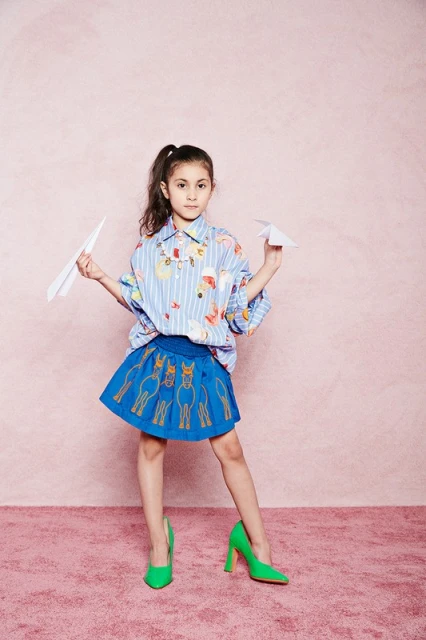 Young girl in patterned shirt and blue skirt poses with paper airplanes on pink background, wearing bright green heels for fashion shoot.