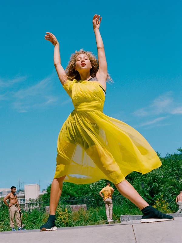 Fashionable woman in a yellow dress with arms raised, standing on concrete in a skate park on a sunny day with people in the background.