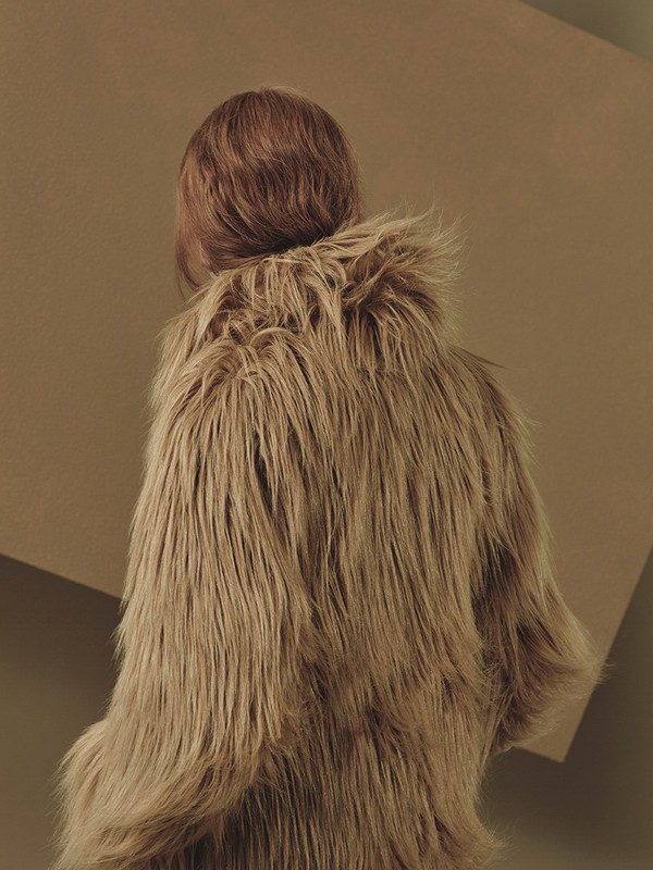 Person from the back wearing a furry brown coat in front of a brown background with a geometric shape, fashion shot.