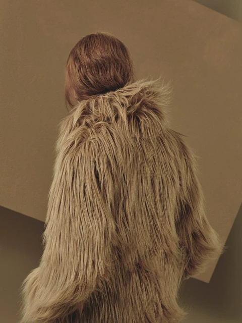 Person from the back wearing a furry brown coat in front of a brown background with a geometric shape, fashion shot.