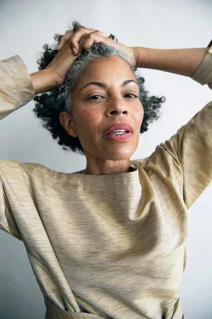 A portrait captures a mature Black woman with hands behind her head, graying curly hair, and a light brown shirt against a white backdrop.