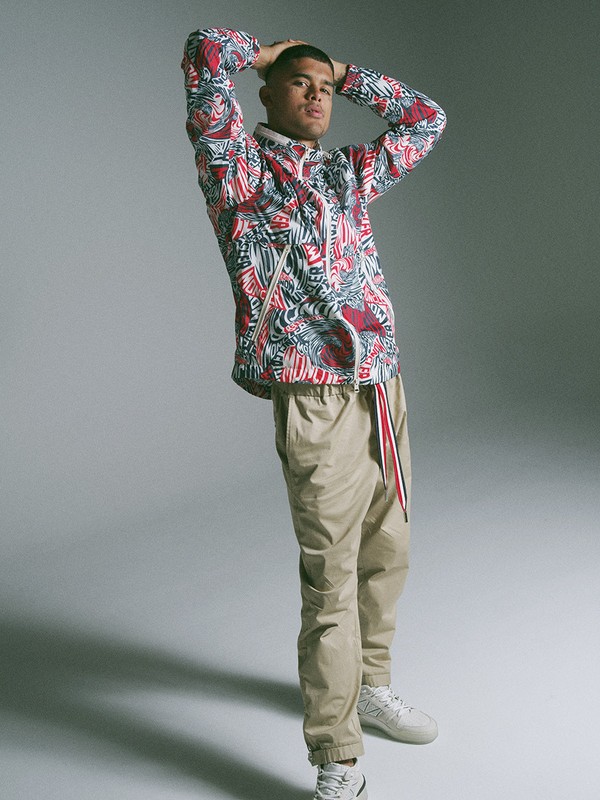 Man wearing a patterned red and white jacket and khaki trousers posing against a grey studio background.