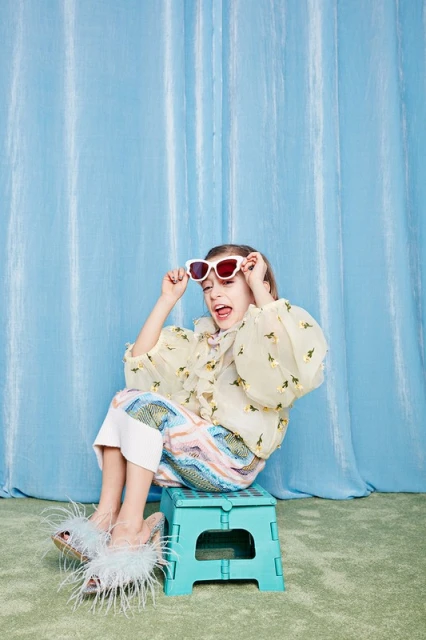 Young girl sits on a teal folding stool wearing sunglasses, a yellow floral blouse, colorful pants and furry slippers against a blue curtain backdrop.