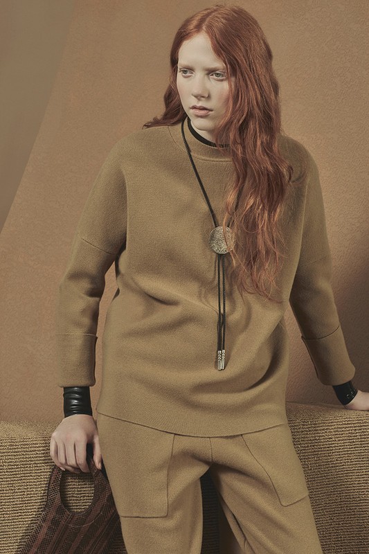 A red-haired woman in a casual brown outfit poses in a photo studio with a neutral background, carrying a brown woven purse.