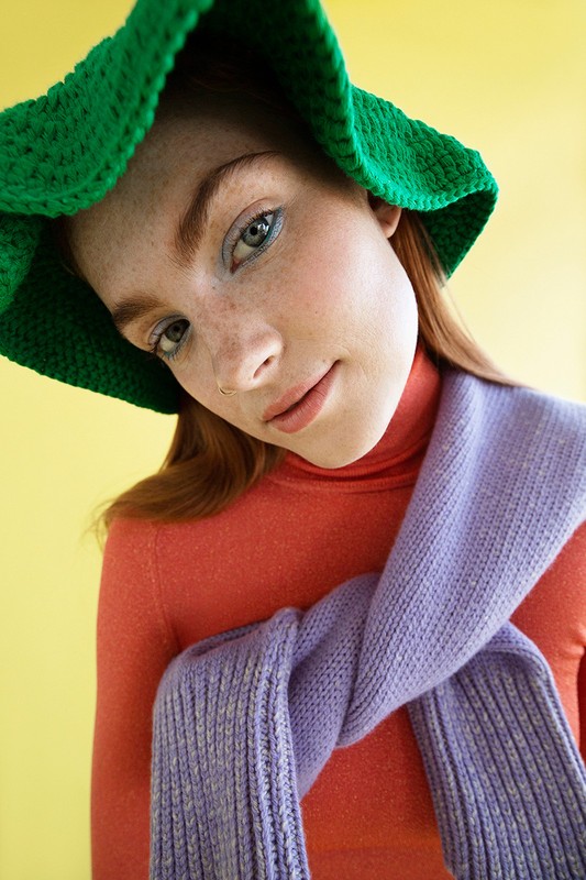 Portrait of a freckled redhead woman wearing a green crochet hat and an orange turtleneck with a lilac sweater draped around her shoulders.