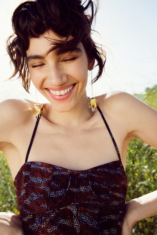 Smiling woman with dark hair and dangling gold earrings wearing a patterned spaghetti-strap dress in outdoor setting. Beauty and fashion.