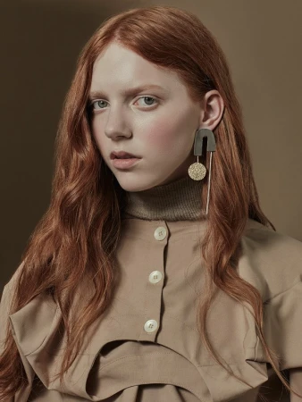Red-haired female model wearing an asymmetrical tan button-up top, turtleneck, and large geometric metallic drop earrings against a brown backdrop