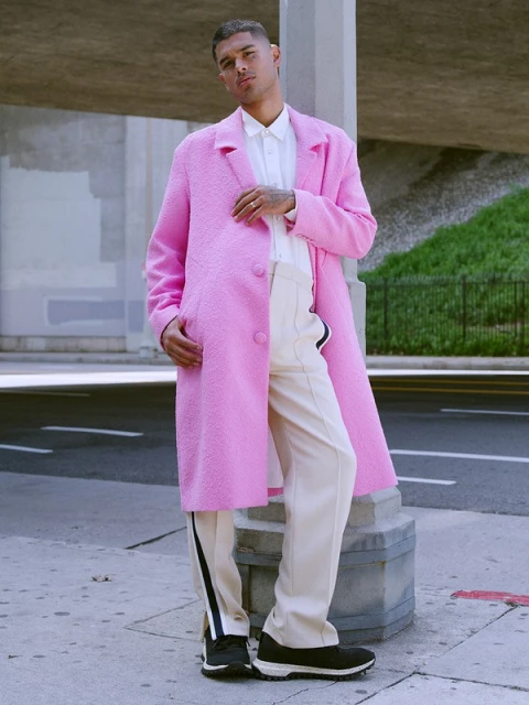 Young adult male stands in a vibrant pink coat, cream pants with a dark stripe, and dark sneakers with light soles in an urban setting.