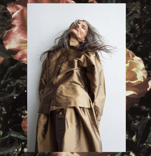 Woman with grey hair in gold satin outfit stands indoors against white wall, dark floral pattern visible in the corners of the image.