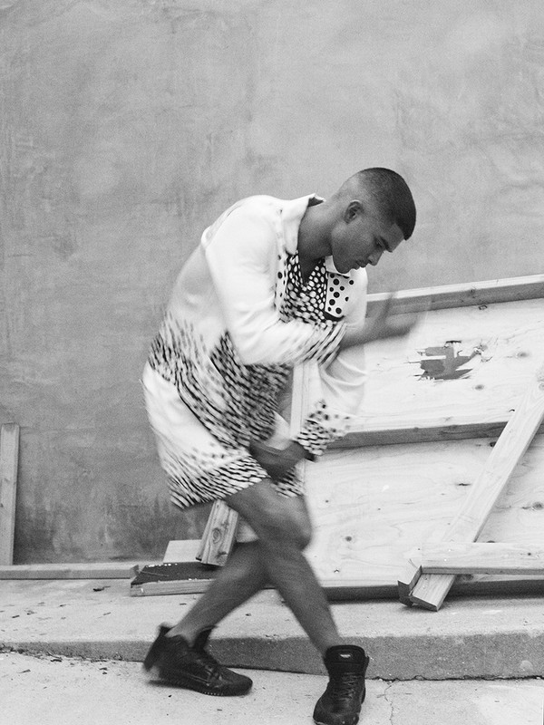 Man in patterned shirt and shorts posing outdoors near wooden panels. Black and white image with casual and fashion elements.