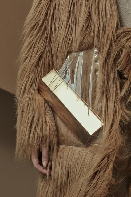 Close-up of a tan faux fur coat with a clear plastic and gold rectangular clutch; studio lighting, neutral tones, and fashion-forward style.