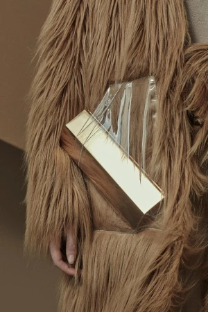 Close-up of a tan faux fur coat with a clear plastic and gold rectangular clutch; studio lighting, neutral tones, and fashion-forward style.