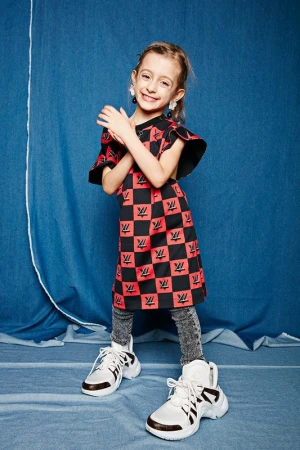 Young girl poses in a red and black dress with white sneakers against a textured blue backdrop in a medium shot, smiling.