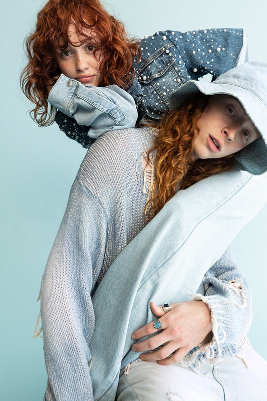 Two red-haired women pose in a studio against a blue backdrop, wearing denim and knitwear in a fashion-forward, relaxed composition.