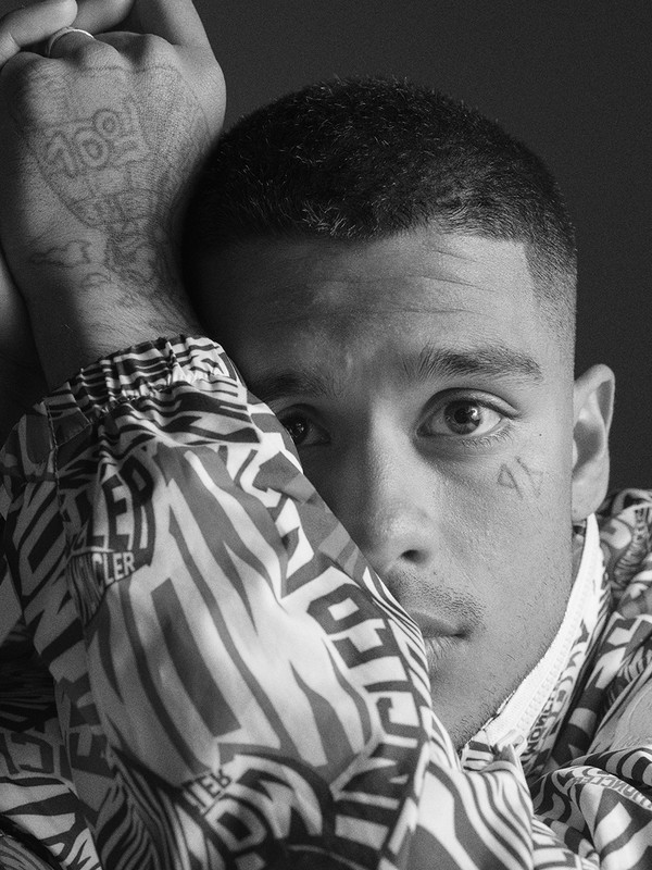Close-up black and white portrait of a young man resting his head on his patterned jacket sleeve, featuring tattoos on his hand and cheek.