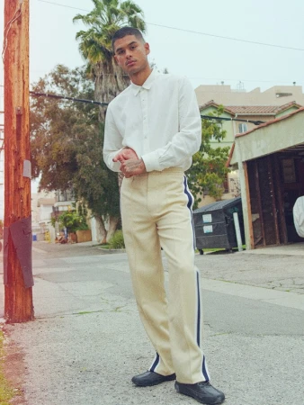 Man in white shirt and tan pants posing on a street with palm tree and buildings in the background under overcast sky. 