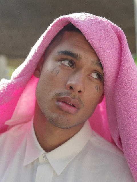 Close-up portrait of a young adult male with a pink textured cloth draped over his head, posing against a blurred background.