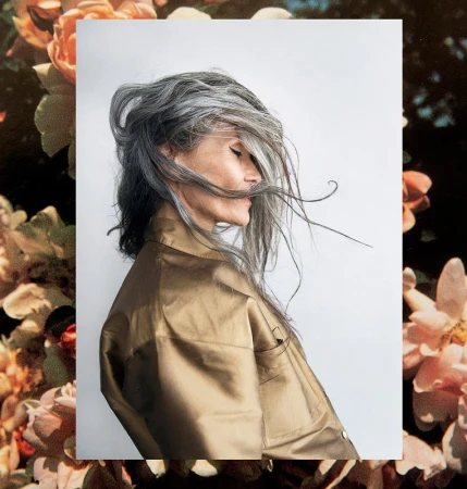 Profile portrait of an elderly woman with long grey hair wearing a metallic bronze shirt against a white seamless backdrop.
