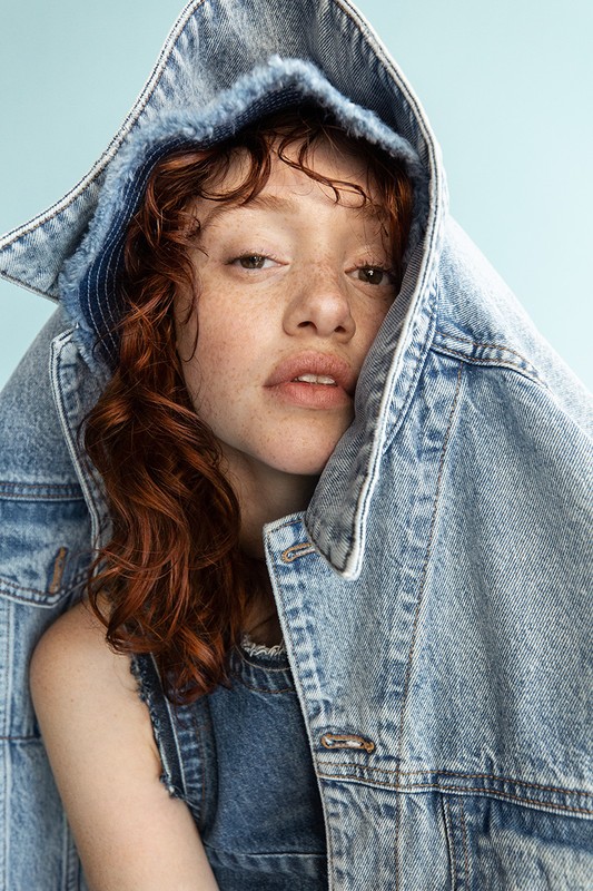 Portrait of a woman with red hair, wearing a denim outfit and jacket over her head against a light blue studio backdrop.